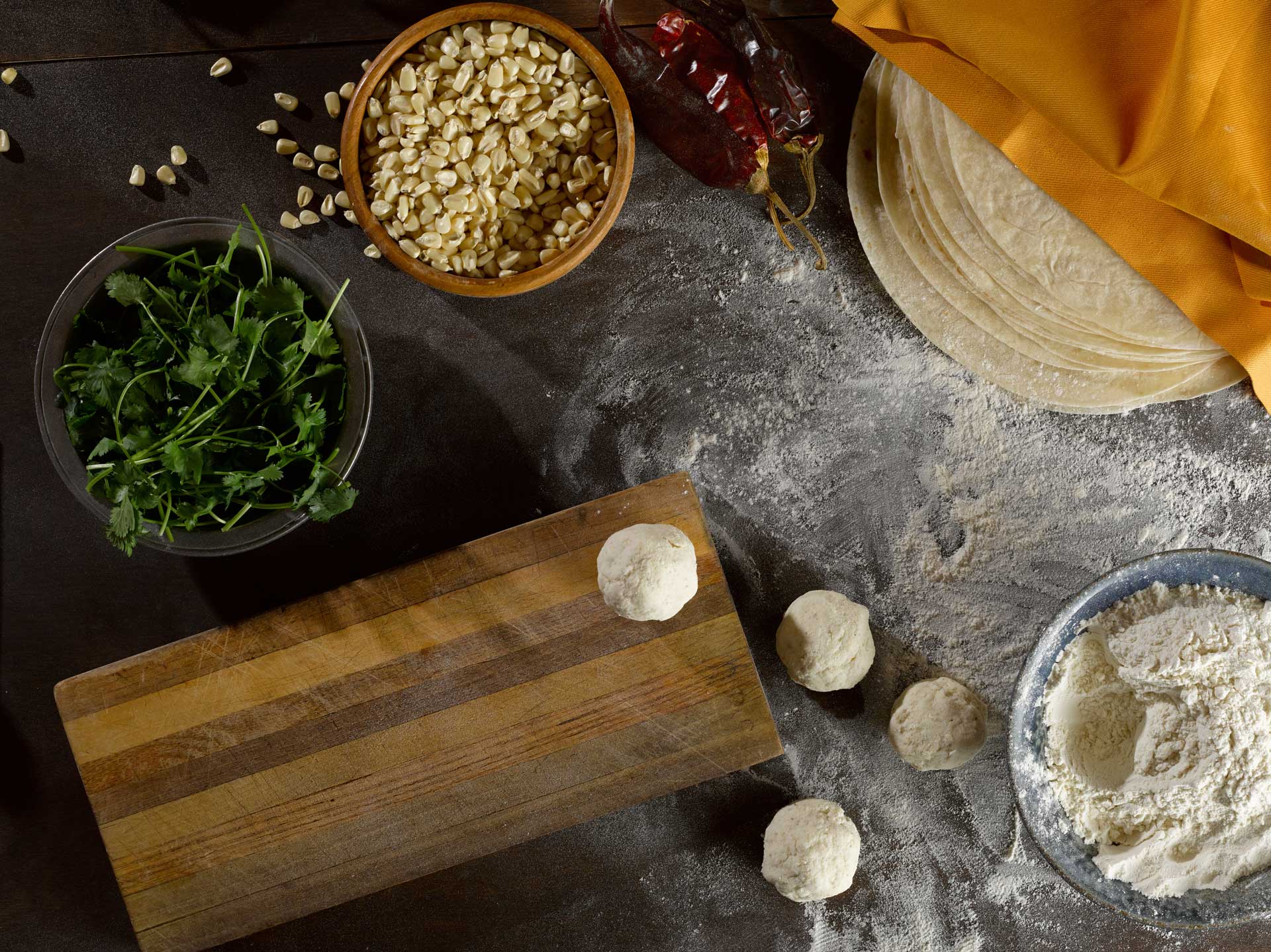 Mama Lola's tortillas, greens, corn, and dough balls rest on a floured board with red chilies and flour