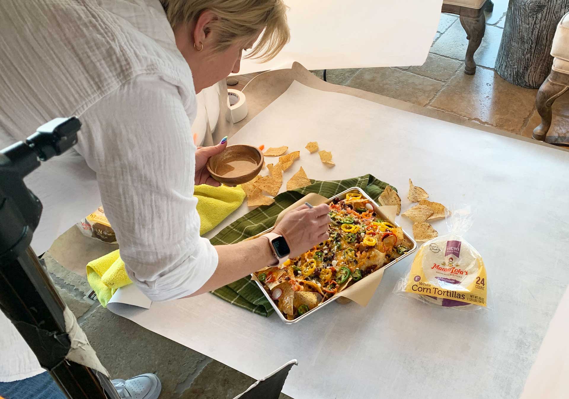 A person arranges toppings on Mama Lola's nachos, surrounded by tortilla chips and food props on a photoshoot set