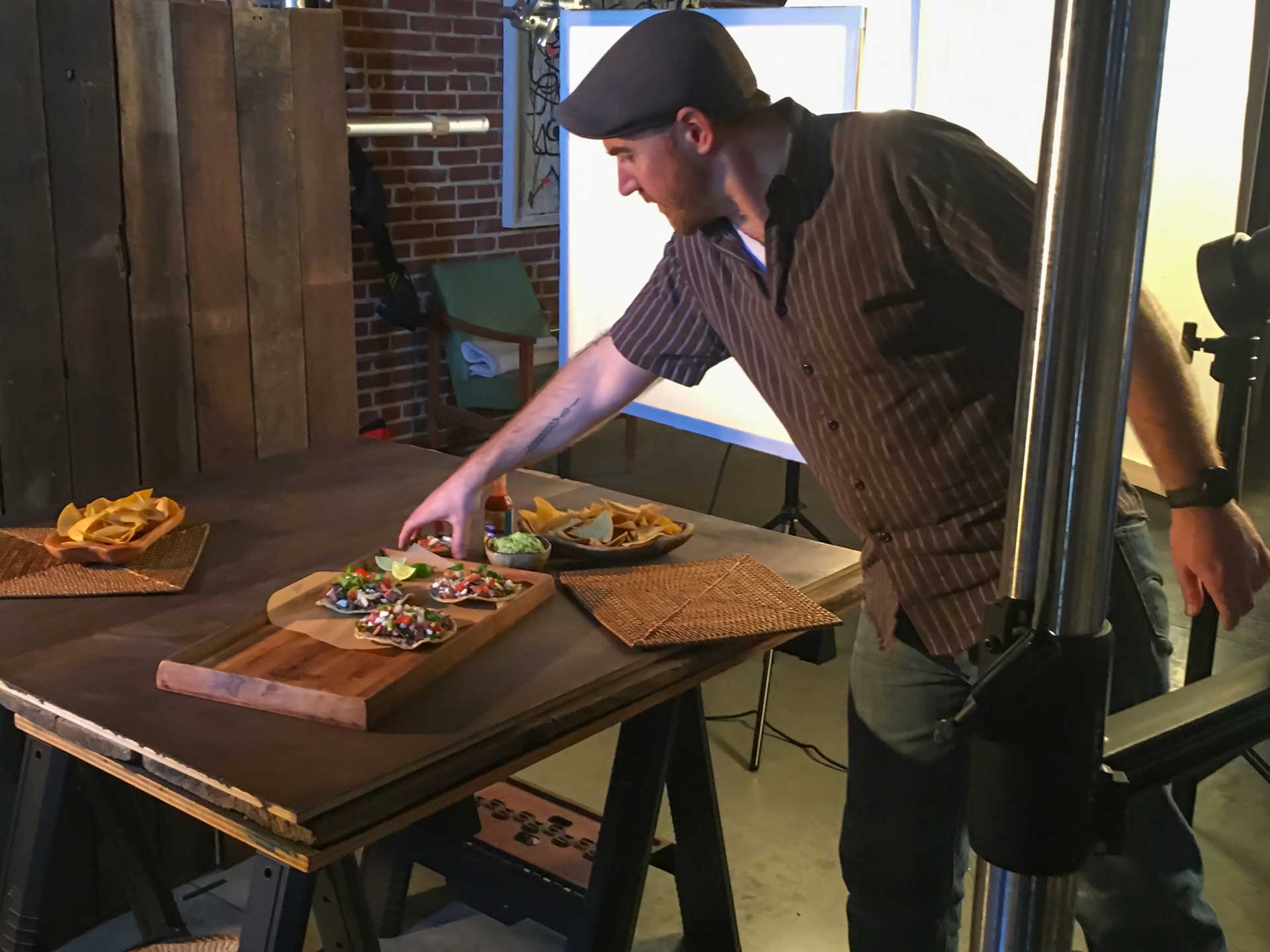 A man arranges food on a wooden table during a photoshoot, with Mama Lola's chips and dip also visible nearby