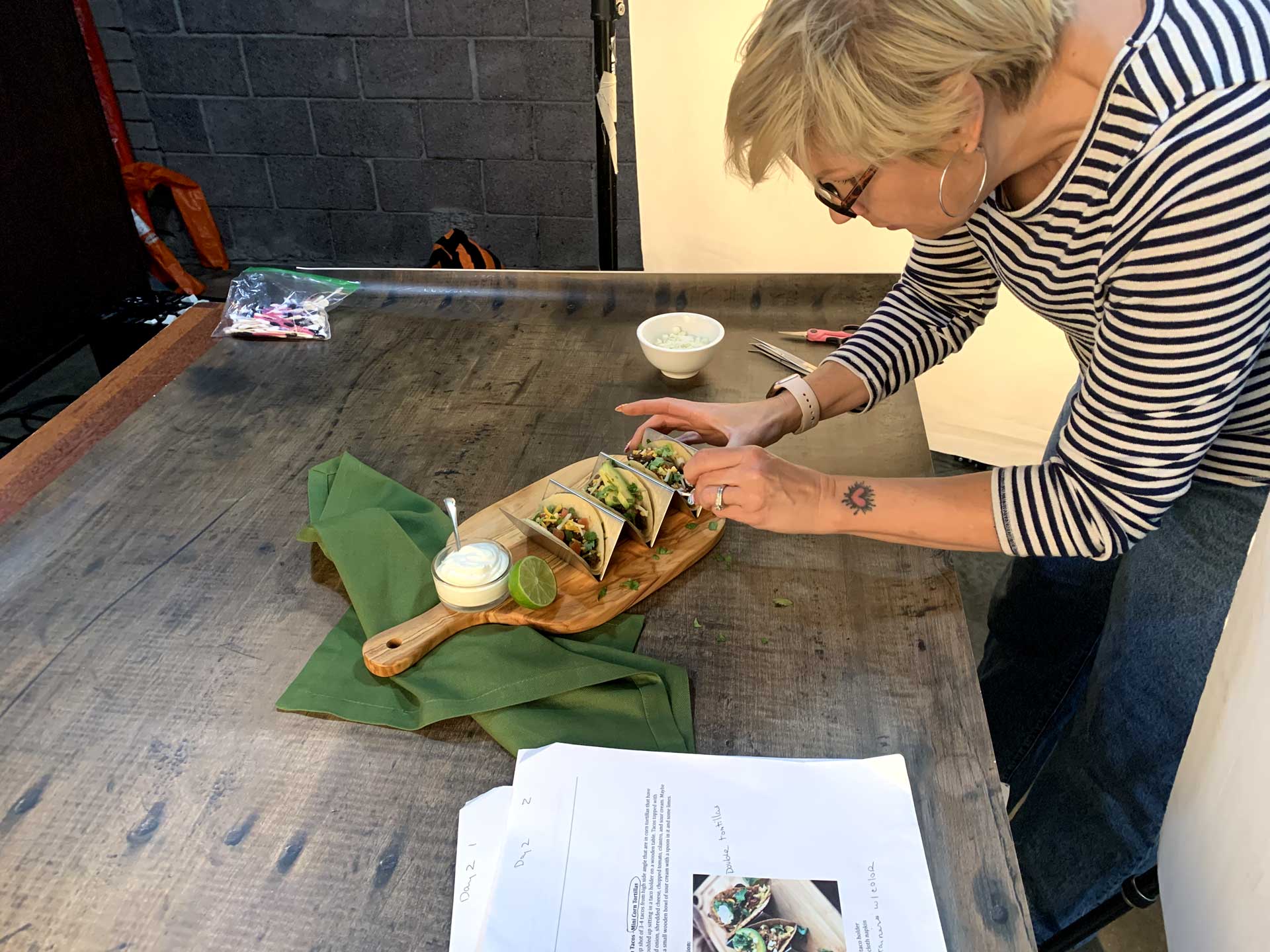 A woman styles Mama Lola's tacos on a wooden board for a food photo shoot, adjusting toppings beside a green napkin