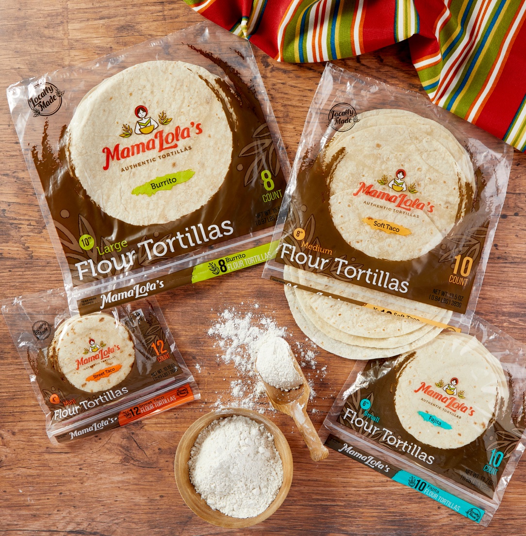 Four packages of Mama Lola's tortillas on wood, with a bowl of flour and a colorful cloth beside them